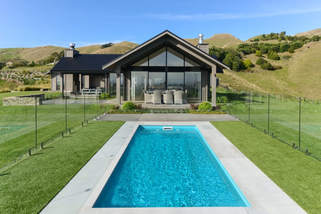 Leisure Pools Esprit in Aquamarine set in a landscaped backyard with light stone paving, glass pool fencing, manicured lawn and a modern gable-roof home with rolling green hills in the background.