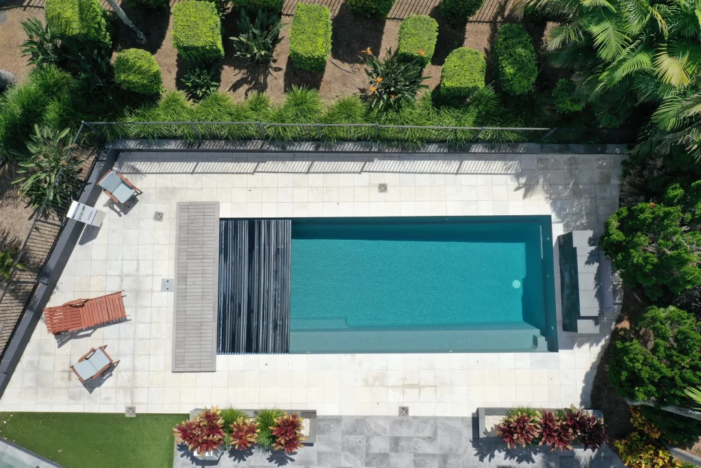 Aerial view of a Leisure Pools Reflection with Auto Cover in Graphite Grey, showing the retractable cover partially stored, full-length bench seat, light stone paving, timber sun loungers, established tropical landscaping and glass pool fencing.