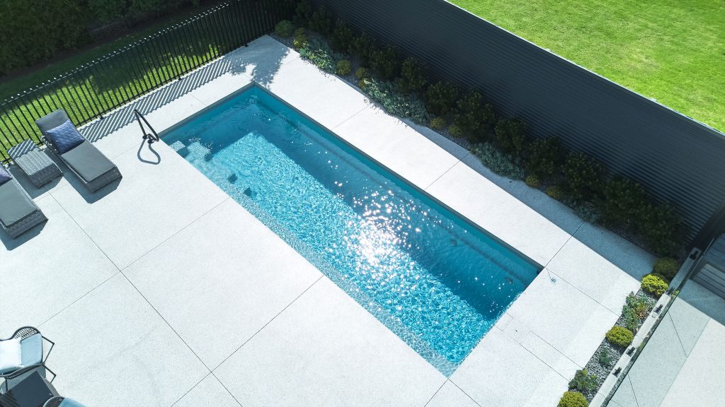 Angled aerial drone photo of a Harmony 8 fibreglass pool in Silver Grey with vivid blue water, viewed from above at an angle showing the pool length, wide large-format light stone paving on three sides, a pool shower and grey sun lounger visible to the upper left, a dark steel louvre boundary fence along the left, frameless glass fencing and low garden planting to the lower right, and a lawn area with established hedging to the upper right