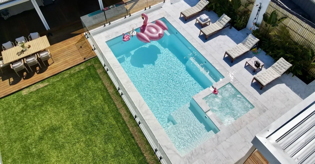 Aerial view of a Leisure Pools Absolute fibreglass pool with spa in Shimmer White finish, surrounded by marble-look paving, timber decking and sun loungers
