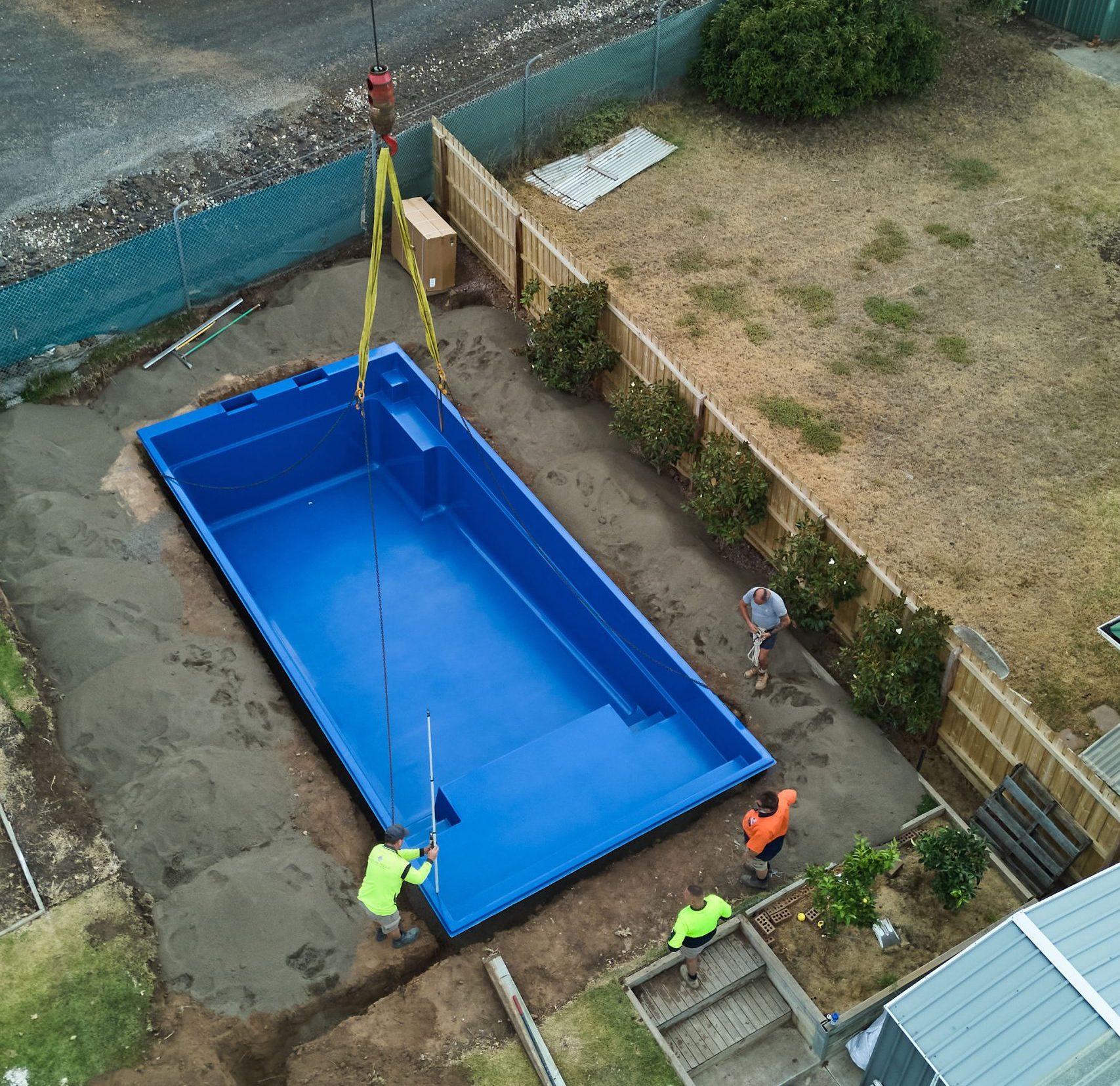 Aerial drone view of a Sapphire Blue Leisure Pools fibreglass shell sitting in an excavated backyard, with built-in steps, bench seat and safety ledge visible, surrounded by exposed earth before paving and landscaping.