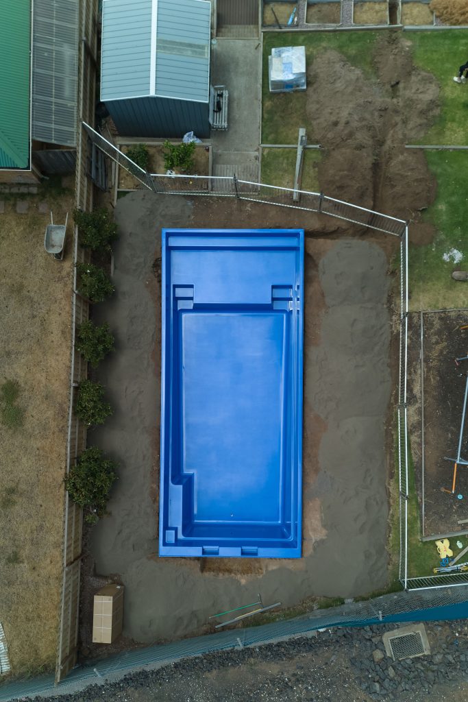 Aerial drone view of a Sapphire Blue Leisure Pools fibreglass shell positioned in an excavated backyard, showing built-in steps, bench seat and safety ledge, with temporary fencing and construction materials surrounding the site.
