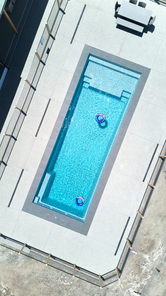 Aerial drone view of a large rectangular fibreglass pool with built-in seating ledge and entry steps, surrounded by light paving, timber and metal pool fencing on all sides, with two Australian flag inflatable rings floating on the water and outdoor furniture visible at the top of frame.