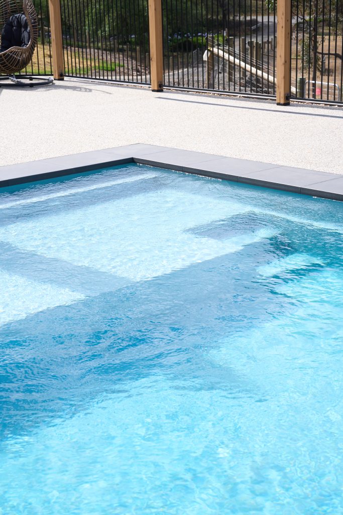 Close-up of a fibreglass pool entry steps and built-in seating ledge through crystal clear water, with dark stone coping, exposed aggregate paving, timber posts and metal pool fencing in the background.