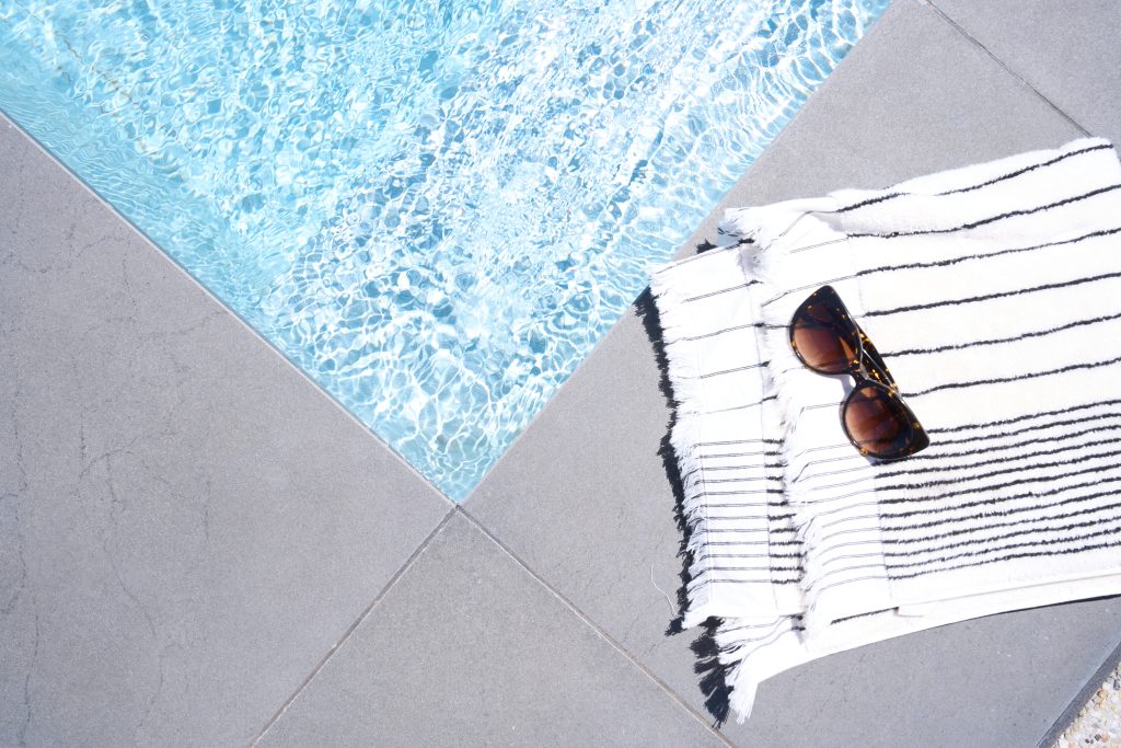 Overhead view of a fibreglass pool edge with dark stone coping, a black and white striped Turkish towel and tortoiseshell sunglasses resting on the poolside paving.
