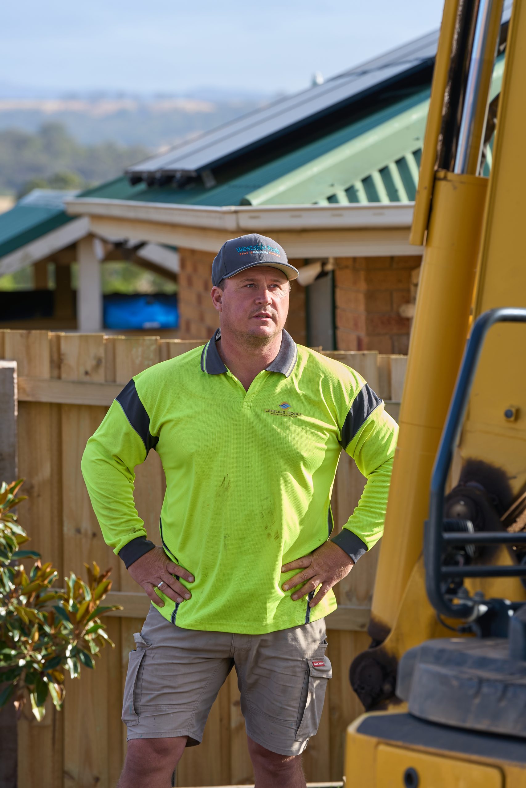 Troy Burrows, owner of Westside Pools, standing on site next to an excavator wearing hi-vis workwear, Leisure Pools polo and branded Westside cap, with a regional Victorian home in the background.