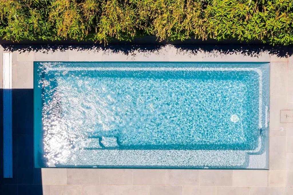 Alt Text (Accessibility) Aerial drone view of a Leisure Pools Harmony 5m in Silver Grey, with full-length bench seat, entry steps and safety ledge visible through sparkling water, surrounded by light stone paving and a mature hedge.